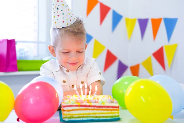 Rubia chico caucásico se sienta pensativa y soñadoramente en la mesa festiva cerca de pastel de arco iris de cumpleaños y pide un deseo. Fondo colorido con globos —  Fotos de Stock