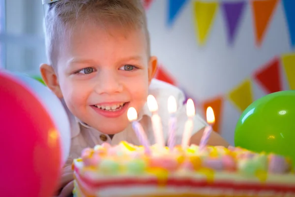 Chico de cumpleaños mirando velas en pastel de arco iris de cumpleaños, pidiendo un deseo antes de soplarlos en la fiesta de cumpleaños. Fondo colorido con globos —  Fotos de Stock