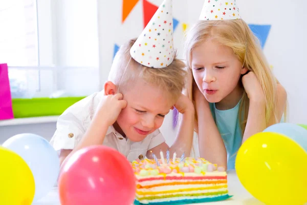 Two blonde caucasian kids boy and girl have fun blowing out candles at birthday rainbow cake with burning candles at birthday party. Stock Image