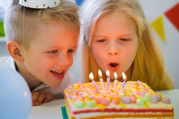 Dos niños caucásicos rubias chico y chica se divierten soplando velas en el pastel de arco iris de cumpleaños con velas ardientes en la fiesta de cumpleaños. — Foto de Stock