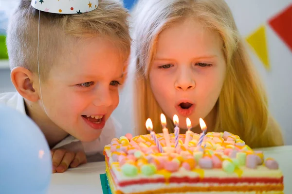 Two blonde caucasian kids boy and girl have fun blowing out candles at birthday rainbow cake with burning candles at birthday party. Royalty Free Stock Photos