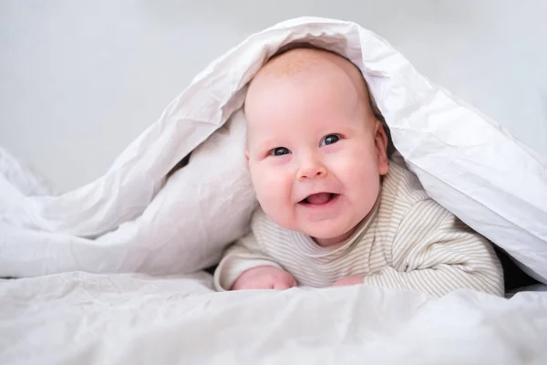 Adorable niño sonriente debajo de una manta blanca en el dormitorio. Niño recién nacido relajándose en la cama. Mañana familiar en casa. Niños recién nacidos — Foto de Stock