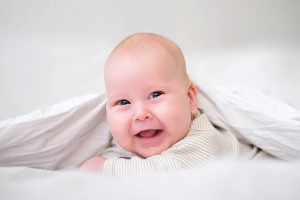 Rozkošný usměvavý chlapeček pod bílou dekou v ložnici. Novorozené dítě odpočívá v posteli. Rodinné ráno doma. Novorozené děti — Stock fotografie