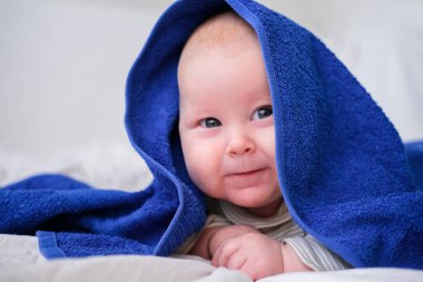 Banyo zamanı. Beyaz yatakta banyo yaptıktan sonra mavi havluyla kameraya bakan sevimli beyaz bebek. Kopyalama alanı..