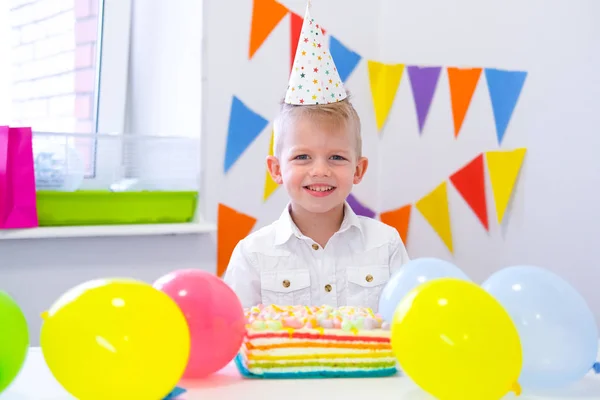 Retrato del chico rubio caucásico sonriendo a la cámara cerca del pastel de arcoíris de cumpleaños. Fondo colorido festivo con globos —  Fotos de Stock