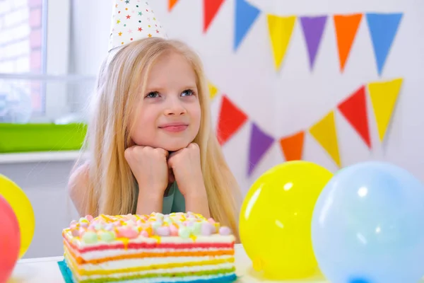 Blonde caucasian girl sits thoughtfully and dreamily at festive table near birthday rainbow cake and makes a wish. Colorful background with balloons — 스톡 사진