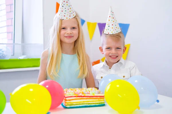 Dos niños caucásicos rubios niño y niña en sombreros de cumpleaños mirando a la cámara y sonriendo en la fiesta de cumpleaños. Fondo colorido con globos y pastel de arco iris de cumpleaños . —  Fotos de Stock