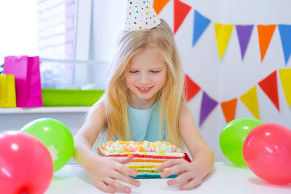 금발의 코카서스 소녀는 생일 무지개 케이크를 보면서 꿈 속에서 웃고 있다. 풍선으로 장식된 화려 한 배경. 생일 파티와 소망의 컨셉 — 스톡 사진
