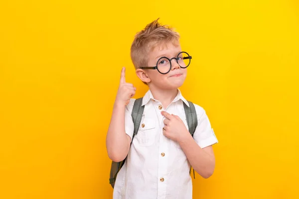 Terug naar school. Portret van een slimme blonde schooljongen met een ronde bril en een tas in een wit shirt. Wijzend omhoog. Gele studio achtergrond. Onderwijs. Kijken en glimlachen naar de camera. — Stockfoto