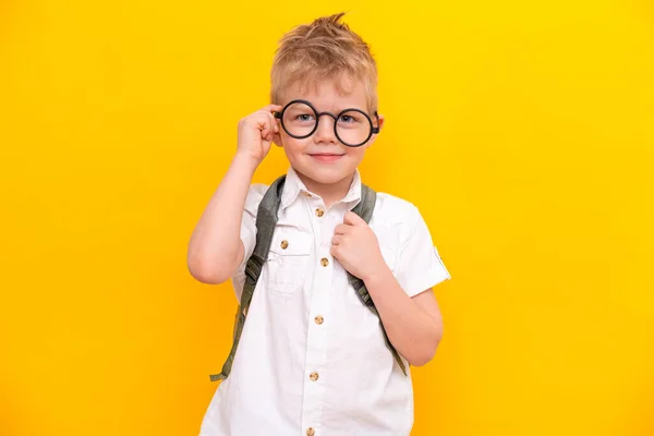 Terug naar school. Portret van een slimme blonde schooljongen met een ronde bril en een tas in een wit shirt. Gele studio achtergrond. Onderwijs. Kijken en glimlachen naar de camera. — Stockfoto