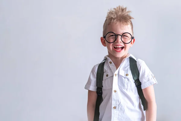 Terug naar school. Portret lachende blonde schooljongen met ronde bril en tas in wit shirt op grijs. Onderwijs. Ik kijk naar de camera. Kopieerruimte — Stockfoto