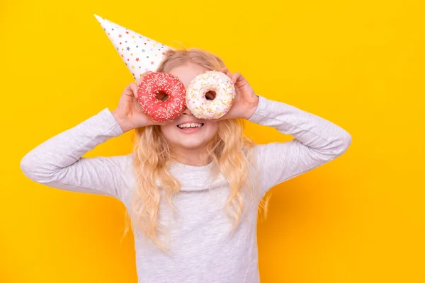 Menina loira bonito em chapéu de aniversário sorrindo, se divertindo e olhando através de dois donuts em seus olhos. Doces. Amarelo estúdio fundo — Fotografia de Stock