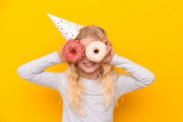 Menina loura alegre louco em chapéu de aniversário sorrindo, se divertindo e olhando através de dois donuts em seus olhos. Doces. Amarelo estúdio fundo — Fotografia de Stock