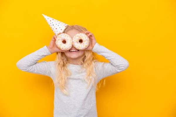 Menina loura alegre louco em chapéu de aniversário sorrindo, se divertindo e olhando através de dois donuts brancos em seus olhos. Doces. Amarelo estúdio fundo — Fotografia de Stock