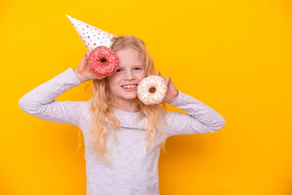 Menina loura alegre louco no chapéu de aniversário sorrindo, se divertindo com dois donuts vermelhos. Feliz aniversário e Sweets. Amarelo estúdio fundo — Fotografia de Stock
