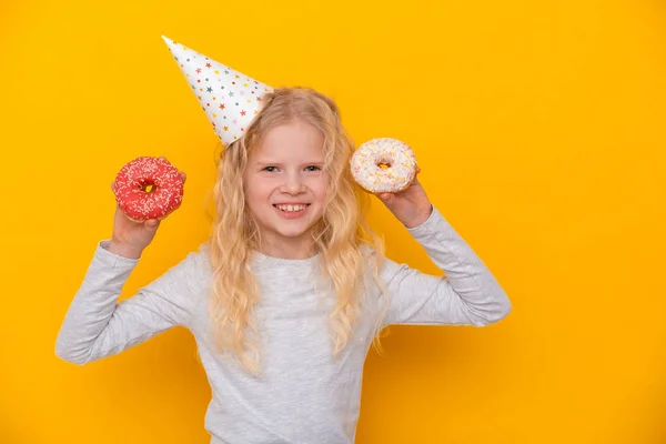 Menina loira bonito em chapéu de aniversário sorrindo, se divertindo com dois donuts envidraçados. Doces. Estúdio amarelo. Espaço de cópia . — Fotografia de Stock