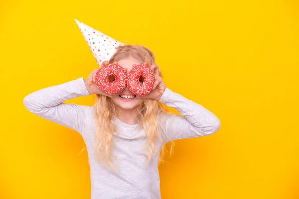 Menina loura alegre louco em chapéu de aniversário sorrindo, se divertindo e olhando através de dois donuts vermelhos em seus olhos. Doces. Amarelo estúdio fundo — Fotografia de Stock