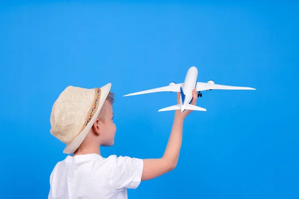 Blonder kaukasischer Junge in Sommermütze und weißer Kleidung mit spielendem Flugzeug in der Hand auf blauem Hintergrund. Reisen und Tourismus — Stockfoto