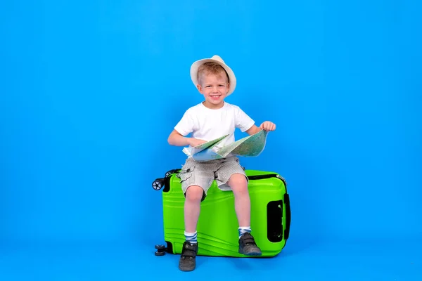 Full length happy blonde little boy summer traveler sitting on green suitcase with map on blue background. travel and tourism, family vacation — Stock Photo, Image