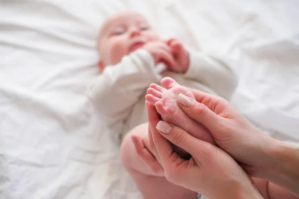 Pies de bebé en manos de madre. Mujer joven caucásica hace masaje para el bebé feliz en la cama blanca en casa. Cuidado de niños, deporte y maternidad feliz . — Foto de Stock