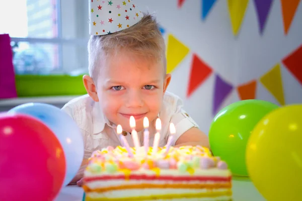 Rubia caucásica niño mirando velas en pastel de arco iris de cumpleaños, pidiendo un deseo antes de soplar hacia fuera en la fiesta de cumpleaños. Fondo colorido con globos —  Fotos de Stock