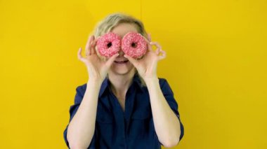 Çılgın neşeli sarışın kız gülümsüyor, eğleniyor ve gözlerindeki iki kırmızı çöreğe bakıyor. Şekerler. Sarı stüdyo geçmişi. 4k görüntü