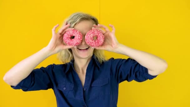Τρελό χαρούμενο ξανθό κορίτσι χαμογελάει, διασκεδάζει και κοιτάζει μέσα από δύο κόκκινα ντόνατς στα μάτια της. Σουίτς. Κίτρινο φόντο στούντιο. 4k πλάνα — Αρχείο Βίντεο