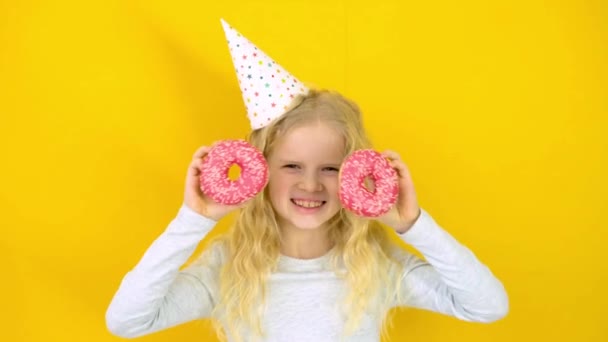 Šílená veselá blondýnka v narozeninovém klobouku se usmívá, baví se a dívá se přes dvě červené koblihy na očích. Sweets. Žluté pozadí studia — Stock video