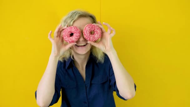 Menina loura alegre louco está sorrindo, se divertindo e olhando através de dois donuts vermelhos em seus olhos. Doces. Estúdio amarelo. Filmagem 4k — Vídeo de Stock