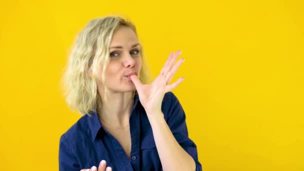 Сумасшедшая весёлая блондинка облизывает пальцы после вкусной еды, изолированной на жёлтом фоне студии. Свитс. 4k кадров — стоковое видео