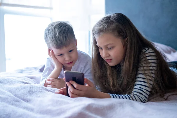 Deux enfants caucasiens garçon et fille jouer à des jeux ou regarder du texte vidéo d'information sur Internet sur les téléphones mobiles à la maison couché dans le lit. Technologie numérique, Enfance moderne — Photo