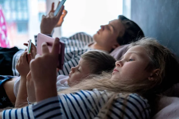 Vue de côté trois enfants caucasiens garçons et filles jouer à des jeux ou regarder du texte vidéo d'information sur Internet sur les téléphones mobiles à la maison couché dans le lit. Technologie numérique, Enfance moderne — Photo