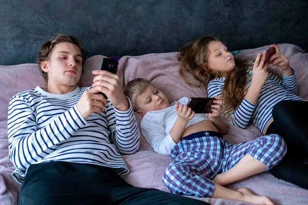 Trzech chłopców i dziewcząt rasy kaukaskiej grających w gry lub szukających informacji wideo w Internecie przez telefony komórkowe w domu leżące w łóżku. Technologia cyfrowa, Nowoczesne dzieciństwo — Zdjęcie stockowe