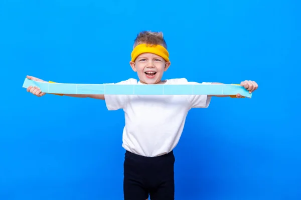 Sportief jongetje met rubberen fitnessband over blauwe studioachtergrond. Gym training. Kindersportman. Activiteit. Sport, gezondheid en energie. Succes — Stockfoto