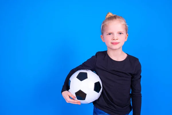 Voetbalfans. Schattig blond meisje in zwart shirt houden voetbal in handen over blauwe studio achtergrond. Kopieerruimte. Voetbalwedstrijd. — Stockfoto