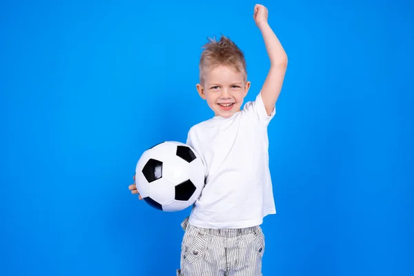Voetbalfans. Vrolijke Kaukasische jongen jongen viert overwinning houden voetbal in handen over blauwe studio achtergrond. Kopieerruimte. Voetbalwedstrijd. — Stockfoto