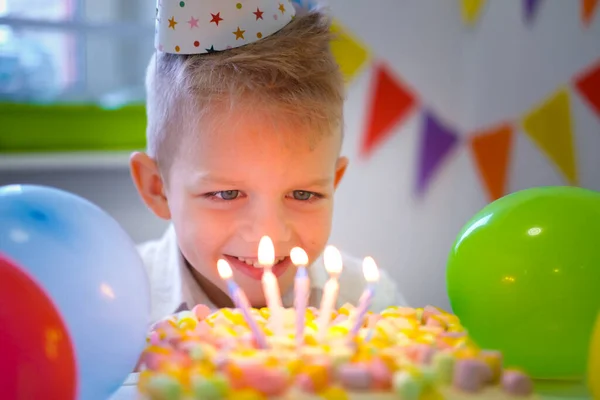 Chico de cumpleaños mirando velas en pastel de arco iris de cumpleaños, pidiendo un deseo antes de soplarlos en la fiesta de cumpleaños. Fondo colorido con globos —  Fotos de Stock