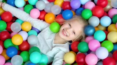Çok renkli plastik topların üzerine uzanmış şirin sarışın bir kız oyun merkezinde büyük kuru bir yüzme havuzunda. Kameraya gülümsüyor. Portre yakın çekim. Oyun odasında eğleniyorum. Boş Etkinlik