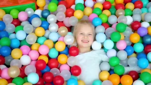 Schattig blond klein meisje liggend op veelkleurige plastic ballen in groot droog peuterbad in speelcentrum. Lachend naar de camera. Portret van dichtbij. Plezier hebben in de speelkamer. Vrijetijdsbesteding — Stockvideo