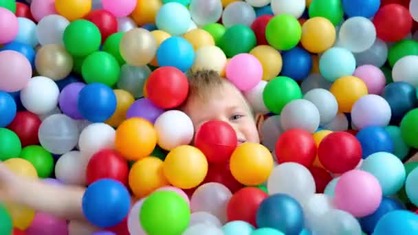 Niño rubio acostado en bolas de plástico multicolores en una gran piscina infantil seca en el centro de juego. Sonriendo a la cámara. Retrato de cerca. Divirtiéndose en la sala de juegos. Actividad de ocio — Vídeos de Stock