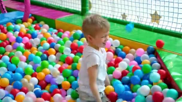 Menino loiro espalha bolas em lados diferentes, deitado em bolas plásticas multicoloridas em grande piscina de remo seco no centro de jogo. A sorrir para a câmara. A divertir-se na sala de jogos. Actividades de lazer — Vídeo de Stock