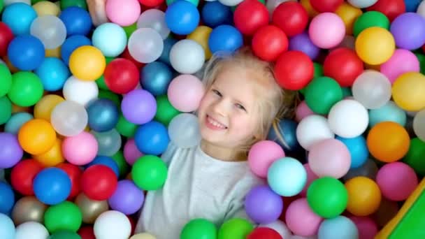 Linda niña rubia acostada en bolas de plástico multicolores en una gran piscina infantil seca en el centro de juego. Sonriendo a la cámara. Retrato de cerca. Divirtiéndose en la sala de juegos. Actividad de ocio — Vídeo de stock