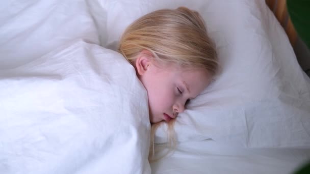 Sova liten blond flicka nära upp i stor säng med vita sängkläder tidigt på morgonen. Högst upp. Barn drömmer. Barndom, lugn, natt .4k film. — Stockvideo