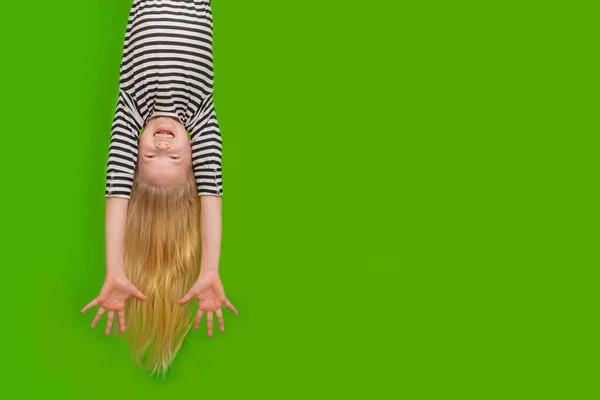 Excited crazy little blonde girl hanging happy upside down hands up over isolated green studio background. Emotion, expression. Copy space for text — Stock Photo, Image