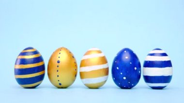 Paskalya yumurtaları yuvarlanıyor, birbirlerini mavi masaya vuruyorlar. Yumurtaların modası geçmiş klasik mavi, beyaz ve altın rengi. Mutlu Paskalyalar. Asgari biçim. 