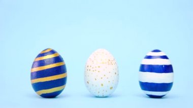 Paskalya yumurtaları yuvarlanıyor, birbirlerini mavi masaya vuruyorlar. Yumurtaların modası geçmiş klasik mavi, beyaz ve altın rengi. Mutlu Paskalyalar. Asgari biçim. 