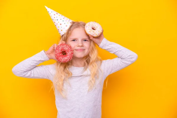Menina loira alegre em chapéu de aniversário sorrindo, se divertindo com dois donuts envidraçados em seus olhos. Doces. Amarelo estúdio fundo — Fotografia de Stock