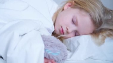 Uyuyan küçük kız rüyasında oyuncak ayıya sarılıyor. Güzel sarışın çocuk sabahın erken saatlerinde beyaz çarşafla yatağında uyuyor. 4k görüntü.