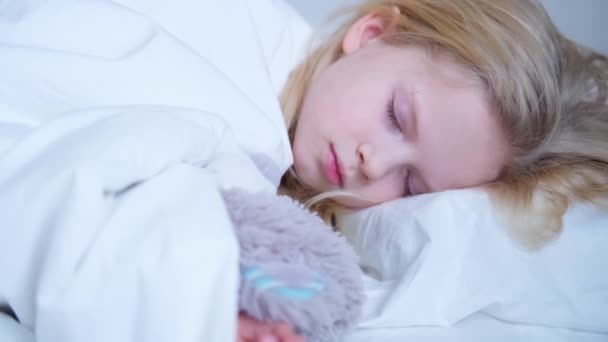 Een slapend meisje knuffelt een teddybeer in een slaapdroom. Mooi blond kind slaapt in haar bed met wit beddengoed vroeg in de ochtend. 4k-beelden. — Stockvideo