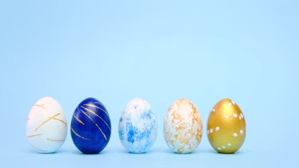 Los huevos de Pascua están rodando, golpeándose unos a otros en la mesa azul. Huevos de moda de color clásico azul, blanco y dorado. Feliz Pascua. Estilo mínimo. — Vídeos de Stock